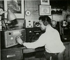 Black and white photo of a man wearing headphones turning a dial on an old radio